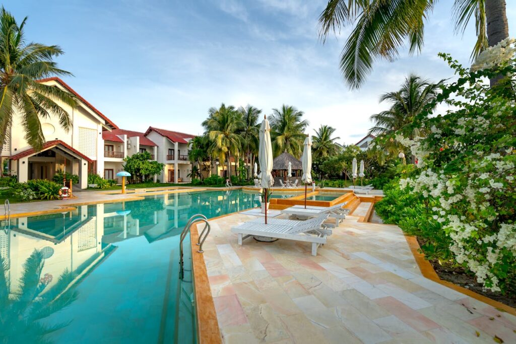 A serene poolside view with sunloungers and villas surrounded by palm trees and lush greenery.