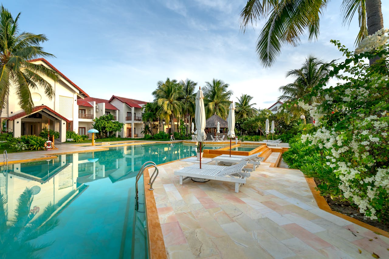 A serene poolside view with sunloungers and villas surrounded by palm trees and lush greenery.